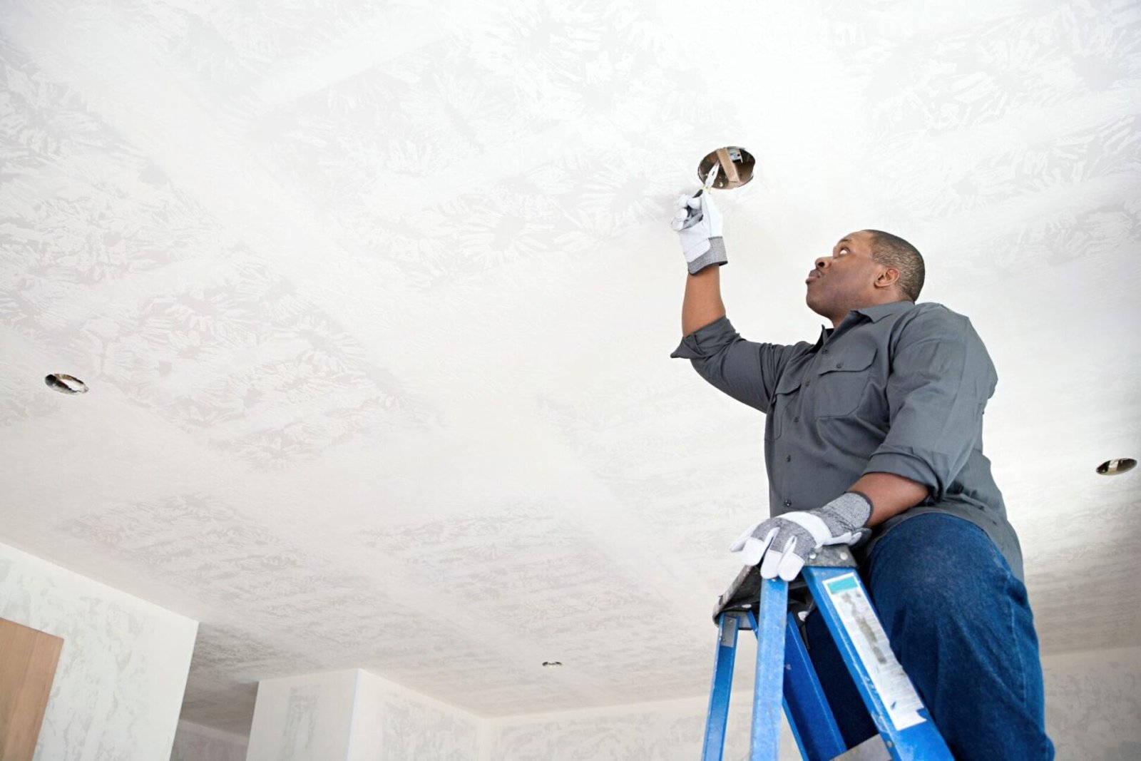 A man on a ladder installs a light fixture in the ceiling of a room. He is wearing gloves and a dark shirt.